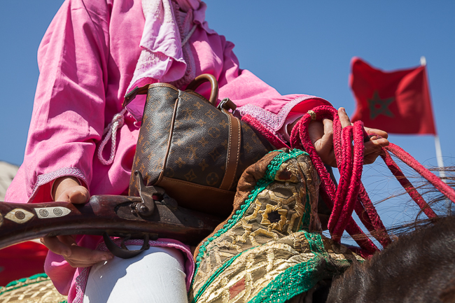 A female Tbourida rider holds her rifle beside a Louis Vuitton-style handbag during a gathering in Morocco.