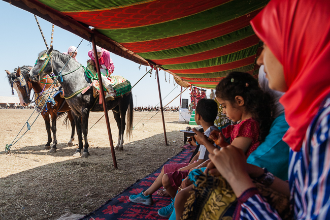 Entre deux cavalcades, des Amazones profitent de la pause et viennent discuter avec le public.
Le public vient majoritairement/principalement des villages voisins. Il est abrité de la chaleur écrasante de l'été par les tentes caïdales traditionnelles. Between two cavalcades, the Amazons take advantage of the break and come to chat with the public. The public comes mainly from nearby villages. They hide from the overwhelming heat of the summer under traditional caidal tents.