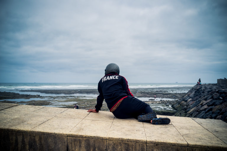 Une personne portant une veste marquée France est assise face à l’océan sous un ciel gris à Casablanca.