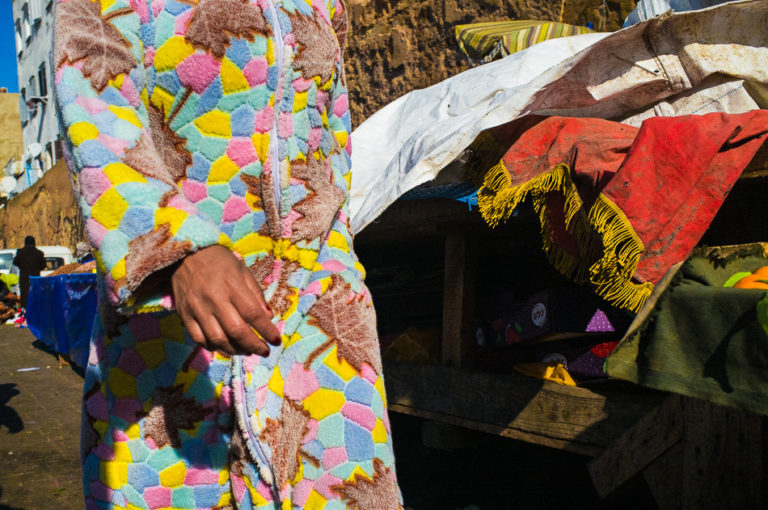 Une personne vêtue d’un habit aux motifs multicolores se tient près de tissus empilés dans un marché de Casablanca.