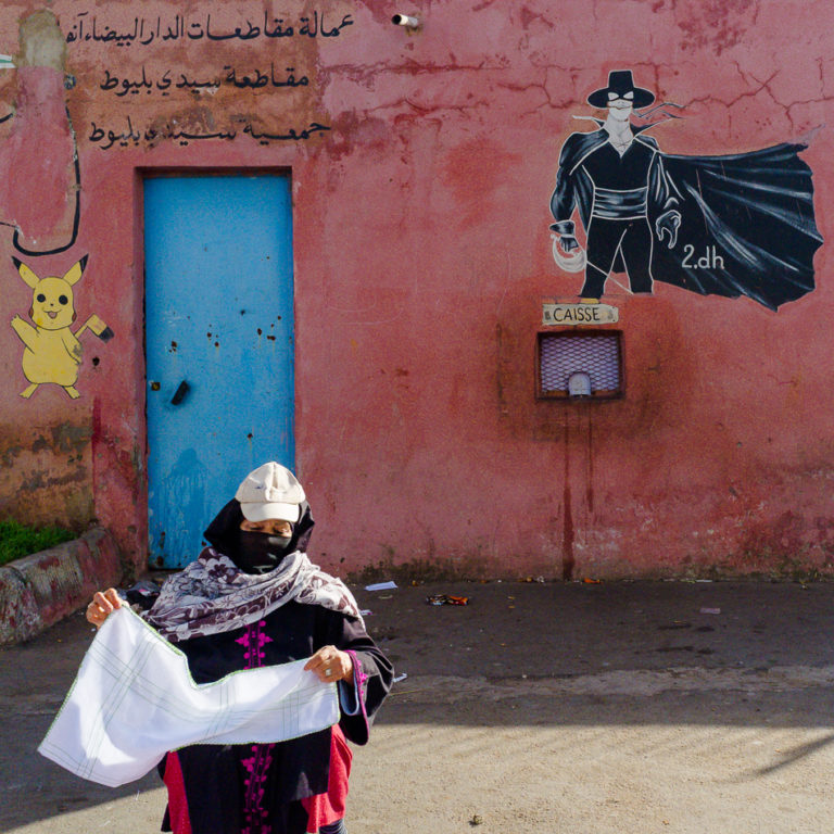 Une femme portant un foulard devant un mur rose peint de personnages et une porte bleue à Casablanca.
