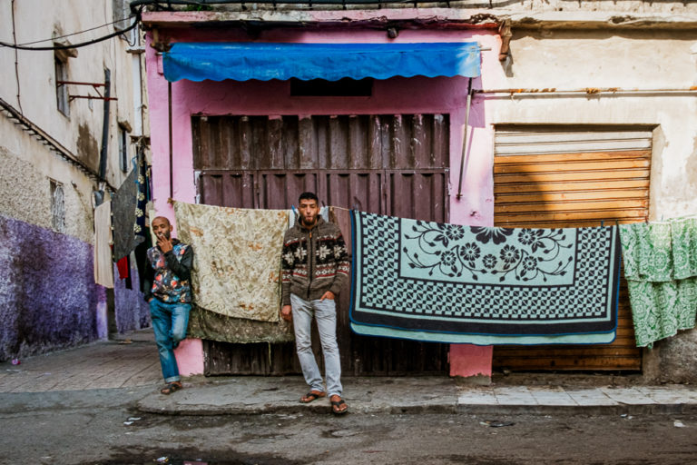 Deux jeunes personnes se tiennent devant des tapis suspendus à la façade colorée d’une rue de Casablanca.