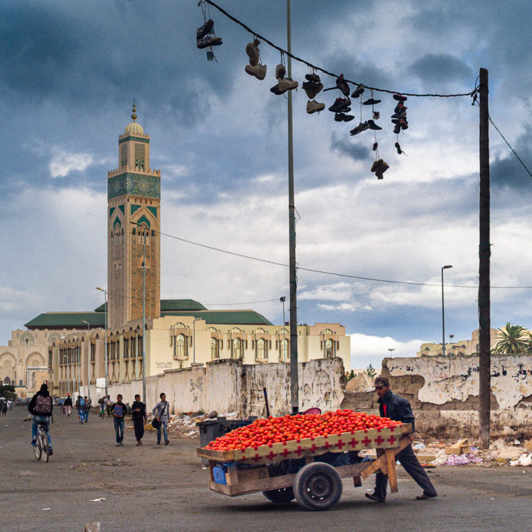 Un vendeur pousse une charrette de tomates devant la mosquée Hassan II, sous des chaussures suspendues à des fils électriques.